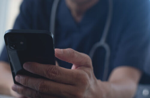 Doctor using mobile smart phone, working on laptop computer in medical workspace office with stethoscope on desk, close up, teleconference, telemedicine, medical research and technology concept