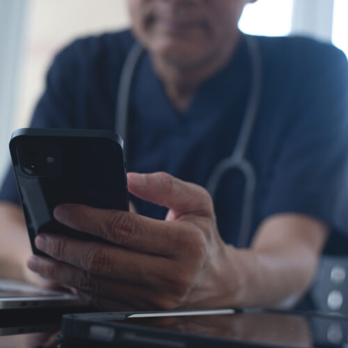 Doctor using mobile smart phone, working on laptop computer in medical workspace office with stethoscope on desk, close up, teleconference, telemedicine, medical research and technology concept