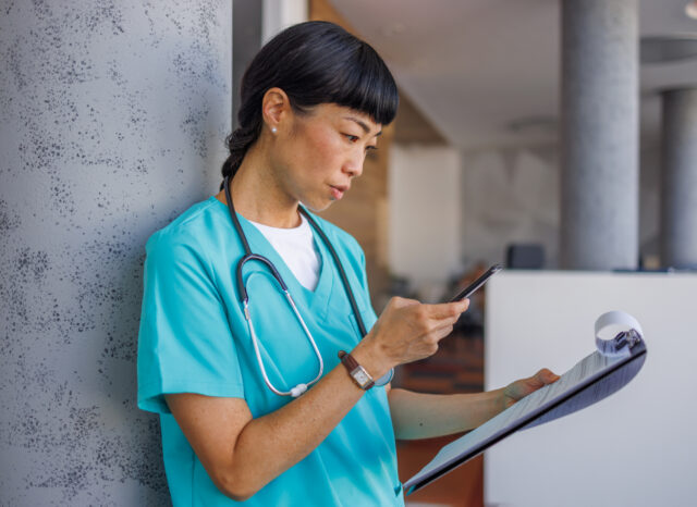 Asian female medical professional is checking patient information on her phone and reading medical report